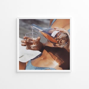 May include: A person pours rosé wine from a bottle into a glass while wearing a blue swimsuit.