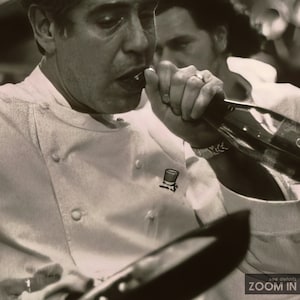 Peut inclure: Une photo en noir et blanc de deux hommes, l'un portant une veste de chef blanche avec un petit logo sur la poitrine gauche, l'autre portant un pull gris. L'homme en veste de chef regarde la cam&eacute;ra et a la bouche ouverte comme s'il parlait. L'homme en pull gris regarde l'homme en veste de chef et a la main sur le menton.
