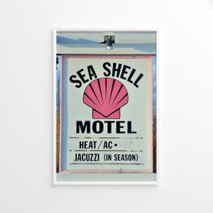 Może przedstawiać: Winietkowy znak motelu z napisem "Sea Shell Motel" czarnymi literami na białym tle. Logo w postaci różowej muszli znajduje się nad tekstem. Znak zawiera również tekst "Ogrzewanie/Klimatyzacja - 8,50 $" i "Jacuzzi (w sezonie)".