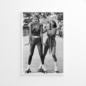Pu&ograve; includere: Due donne con abiti in stile anni '80 stanno pattinando su una strada cittadina. Una donna indossa una tuta nera con una cintura e l'altra indossa un vestito corto con volant. Entrambe le donne indossano pattini bianchi.