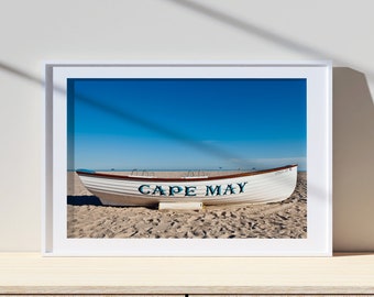 Może przedstawiać: Biała łódź ratunkowa z napisem "CAPE MAY" namalowanym na boku, stojąca na piaszczystej plaży z błękitnym niebem w tle.