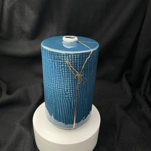 Op de afbeelding: Een cilindrische blauwe keramische vaas met een gestructureerd ruitpatroon op het bovenste gedeelte en een gladde afwerking aan de onderkant. De vaas vertoont een opvallende goudkleurige reparatielijn die verticaal over het oppervlak loopt.