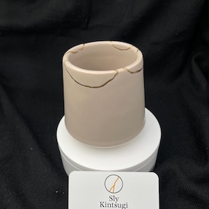 Könnte beinhalten: Beige Keramikbecher mit goldfarbenen Kintsugi-Reparaturlinien. Der zylindrische Becher steht auf einem weißen Podest. Eine Visitenkarte mit dem Sly Kintsugi-Logo und Kontaktinformationen liegt davor.