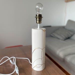 Puede incluir: Lámpara de mesa de cerámica blanca con detalles dorados y una bombilla transparente. La lámpara tiene una base cilíndrica con un acabado craquelado y líneas dorées.