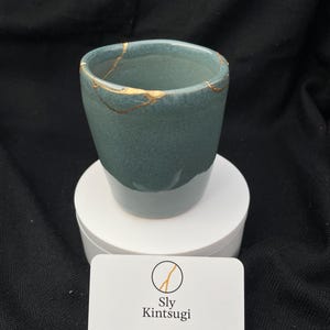 Può includere: Una tazza in ceramica color teal con linee di riparazione kintsugi dorate. La tazza è su un piedistallo bianco, con un biglietto da visita davanti. La carta ha il testo "Sly Kintsugi" e informazioni di contatto.