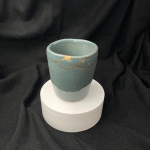 Könnte beinhalten: Ein Keramikbecher mit zweifarbigem Design, bestehend aus einem gedeckten blaugrünen oberen Teil und einer helleren graublauen Basis. Der obere Bereich weist einen deutlichen Riss auf, der mit goldfarbenem Material repariert wurde und ein Muster bildet.