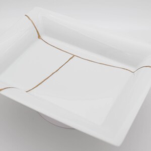 Op de afbeelding: Witte rechthoekige serveerschaal met gouden lijnen. De schaal heeft een verhoogde rand en een licht gebogen basis.
