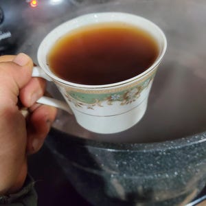 May include: A white teacup with a green and gold floral design, filled with dark brown tea. The teacup is being held by a hand over a black pot.