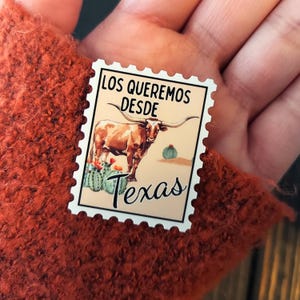 May include: A rectangular sticker resembling a postage stamp, featuring a longhorn steer, cacti, and the text "LOS QUEREMOS DESDE Texas". The stamp has a cream background and is held against a textured orange surface.