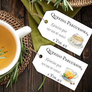 May include: Two white gift tags with text in Spanish, "QUERIDA PRECURSORA" and "QUERIDO PRECURSOR", with watercolor illustrations of teacups. A white teacup filled with tea and rosemary sprigs sits on a wooden surface.