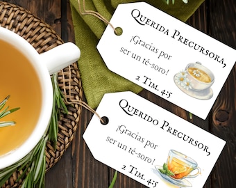 May include: Two white gift tags with text in Spanish, "QUERIDA PRECURSORA" and "QUERIDO PRECURSOR", with watercolor illustrations of teacups. A white teacup filled with tea and rosemary sprigs sits on a wooden surface.