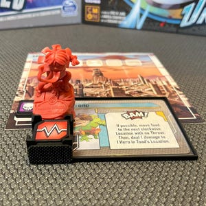 Puede incluir: Una figura de plástico roja de una niña con coletas, de pie sobre una base roja con un símbolo de calavera blanco y negro. La figura está en un tablero de juego con una carta que dice "BAM! Si es posible, mueve a Toad a la siguiente ubicación en el sentido de las agujas del reloj sin Amenaza. Luego, inflige 1 daño a 1 Héroe en la ubicación de Toad."