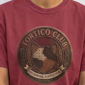Può includere: Una maglietta bordeaux con un emblema circolare con la scritta "TORTICO CLUB" e "PATCHWORK PERFECTION ORIGINAL". L'emblema presenta un disegno di volto di gatto con un motivo patchwork marrone e bianco.