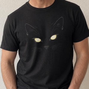 May include: Black t-shirt with a graphic of a cat's face, showing yellow glowing eyes. The design includes the cat's ears, nose, and whiskers. The t-shirt is made of a soft material, suitable for casual wear.