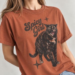 Peut inclure: Un t-shirt à manches courtes de couleur rouille arborant l'illustration d'un chat écaille de tortue la gueule ouverte. Le texte "Spicy Girl" est imprimé dans une police sombre et stylisée au-dessus du chat, avec des étoiles à quatre branches.