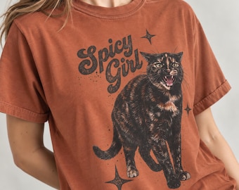 Puede incluir: Una camiseta de manga corta color óxido con el gráfico de un gato carey con la boca abierta. El texto "Spicy Girl" está impreso en una fuente oscura y estilizada sobre el gato, acompañado de pequeños iconos de estrellas de cuatro puntas.