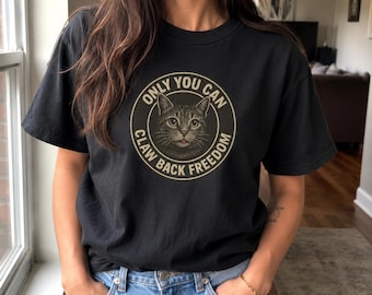 May include: Black t-shirt with a circular graphic featuring a cat's face and the text "ONLY YOU CAN CLAW BACK FREEDOM". The cat graphic is in a vintage style. The shirt is paired with blue jeans.
