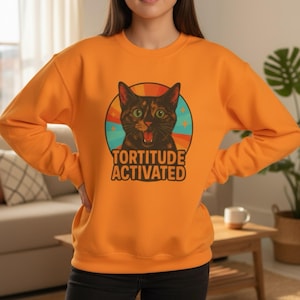 Può includere: Felpa arancione con grafica di gatto e la scritta "TORTITUDE ACTIVATED". Il gatto è marrone e nero con occhi verdi, su un design a cerchio arcobaleno retrò. Un top casual e confortevole.