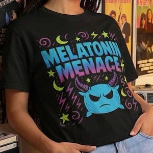 Könnte beinhalten: Schwarzes T-Shirt mit dem Text "MELATONIN MENACE" in einem Farbverlauf aus Blau, Pink und Lila. Das Design umfasst Sterne, Monde, Blitze und ein blaues Monster mit Hörnern.