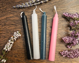 May include: Four small taper candles in black, white, dark blue, and pink. The candles are arranged in a row on a wooden surface.