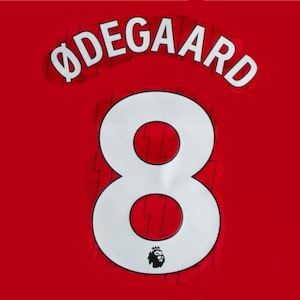 May include: Red jersey with white lettering and numbers. The name "ØDEGAARD" is arched across the top, with the number "8" below. The Premier League logo is at the bottom. The jersey is a solid red color.