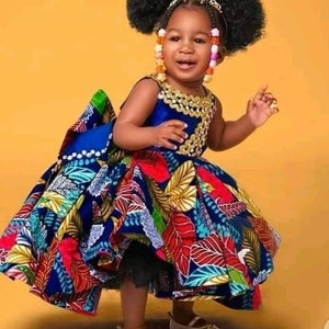 Könnte beinhalten: Ein junges Mädchen trägt ein farbenfrohes, gemustertes Kleid mit einem blauen Oberteil und einer goldenen Halskette. Ihre Haare sind zu zwei Afro-Pferdeschwänzen mit bunten Perlen gestylt.
