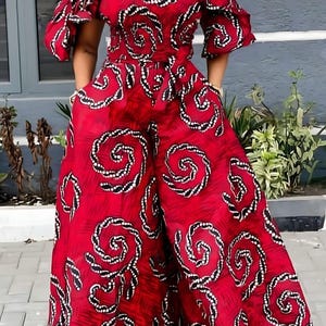 May include: A red jumpsuit featuring an off-the-shoulder neckline, short ruffled sleeves, and wide-leg trousers. The fabric displays a repeating pattern of black and white circular spiral motifs throughout the entire garment.