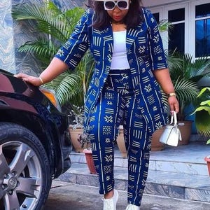 May include: A navy blue and white patterned trouser suit with a matching blazer and trousers. The outfit is accessorised with a light pink hat, white sunglasses, and white trainers. A small white handbag completes the look.