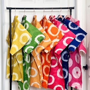 May include: A clothing rack displays a collection of colorful shirts. Each shirt features a large white circle design on a solid background. The shirts are in various colors, including yellow, green, orange, pink, and blue.
