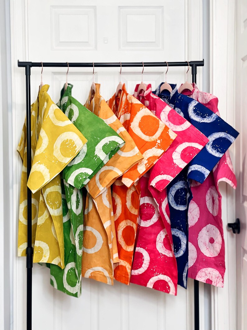 May include: A clothing rack displays a collection of colorful shirts. Each shirt features a large white circle design on a solid background. The shirts are in various colors, including yellow, green, orange, pink, and blue.