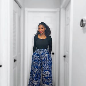 May include: A woman wearing a black long-sleeve top and wide-leg pants. The pants are a vibrant blue with a white floral pattern. The outfit is styled with a simple necklace and sandals.