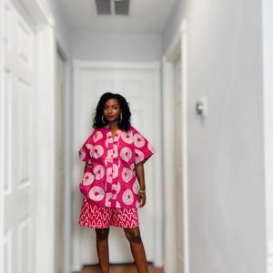 May include: A two-piece outfit featuring a short-sleeved, button-down shirt and matching shorts. The set is a vibrant pink with a white circular pattern on the shirt and a red and white geometric design on the shorts. The outfit is paired with red high-heeled sandals.