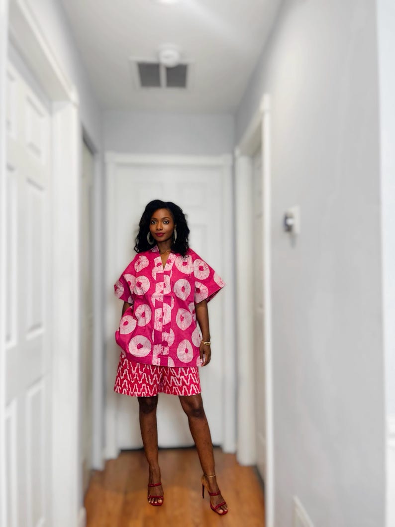 May include: A two-piece outfit featuring a short-sleeved, button-down shirt and matching shorts. The set is a vibrant pink with a white circular pattern on the shirt and a red and white geometric design on the shorts. The outfit is paired with red high-heeled sandals.
