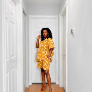 May include: A mustard-yellow two-piece outfit with a white abstract pattern. The outfit consists of a short-sleeved button-down shirt and matching shorts. The person is wearing red high-heeled sandals.