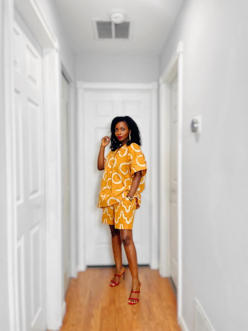 May include: A mustard-yellow two-piece outfit with a white abstract pattern. The outfit consists of a short-sleeved button-down shirt and matching shorts. The person is wearing red high-heeled sandals.