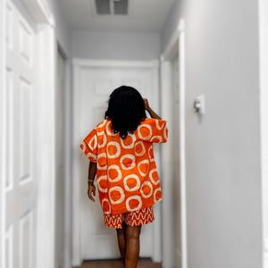 May include: An orange two-piece outfit with white circular patterns on the top and a matching patterned skirt. The outfit is worn by a person walking down a hallway with white doors and walls.