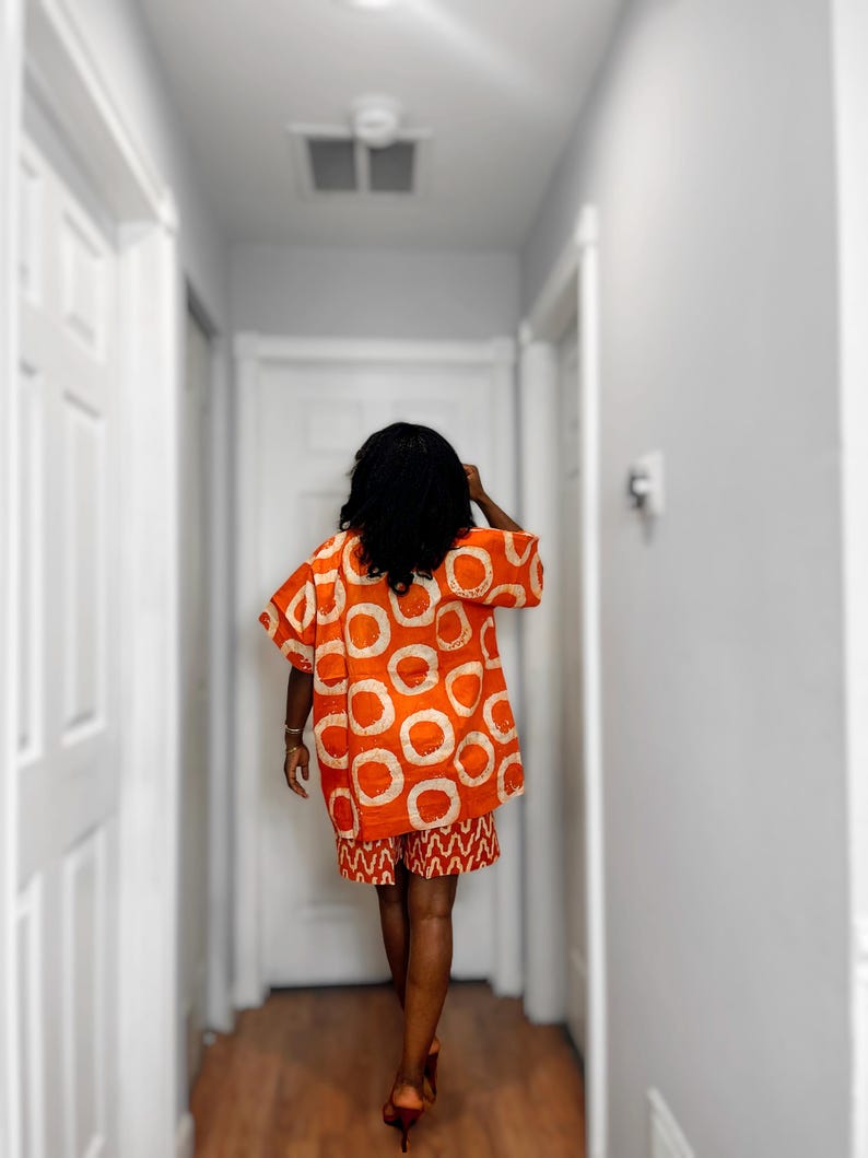 May include: An orange two-piece outfit with white circular patterns on the top and a matching patterned skirt. The outfit is worn by a person walking down a hallway with white doors and walls.