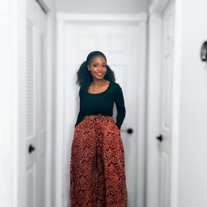 May include: A woman wearing a black long-sleeve top and wide-leg pants. The pants have a red and brown pattern. The woman is smiling and standing in a hallway with white doors.