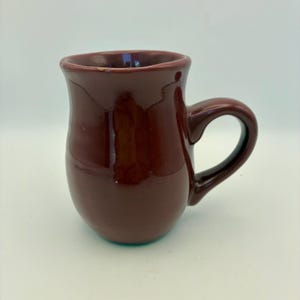 Pode incluir: Uma caneca de cerâmica marrom com um corpo arredondado e uma alça curva. A caneca tem um acabamento brilhante e uma borda ligeiramente alargada. A alça tem um formato de laço simples e a caneca parece ser feita à mão.