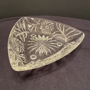 Puede incluir: Un plato de servir triangular de vidrio transparente con un diseño de vidrio cortado. El plato tiene un borde festoneado y tres pequeños pies.