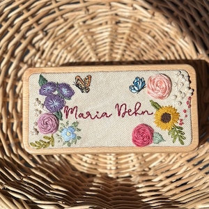 May include: A rectangular wooden plaque with embroidered floral and butterfly details. The design includes purple morning glories, pink roses, a yellow sunflower, and a blue butterfly. The name "Maria Dehn" is embroidered in red script.