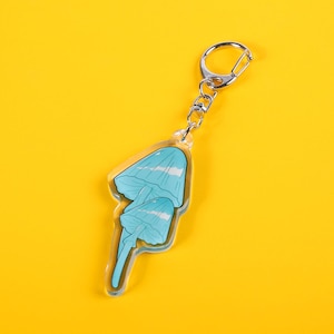 Op de afbeelding: Een heldere acryl sleutelhanger met een lichtblauw paddenstoelontwerp. De sleutelhanger heeft een zilverkleurige metalen sluiting en ketting. Het paddenstoelontwerp heeft twee paddenstoelen met blauwe doppen en stelen. De sleutelhanger staat op een gele achtergrond.