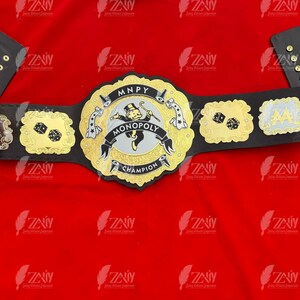 May include: A black and gold championship belt with a Monopoly themed center plate. The center plate features the Monopoly Man character and the text "MNPY MONOPOLY CHAMPION".