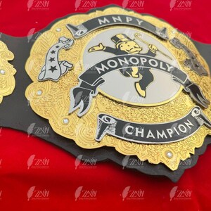 May include: A gold and black championship belt with a Monopoly theme. The belt features a silver center plate with a Monopoly Man figure, the words "MONOPOLY" and "CHAMPION", and a dice design on the side.