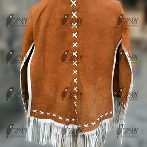 May include: A brown suede jacket with white fringe and trim. The back of the jacket features white cross-stitch detailing down the center and along the bottom edge. The jacket has a classic, rustic design.