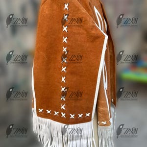 May include: A brown suede jacket with white fringe and accents. The jacket features white stitching and trim, with a fringe detail along the bottom. The jacket is displayed on a mannequin.