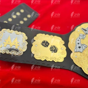 May include: A black leather championship belt with gold and silver accents. The belt features a large, ornate silver plate with the number "4" in the center. The belt also has a smaller gold plate with two dice and a larger gold plate with a stylized image of a person holding a playing card.