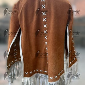 May include: A brown suede jacket with white fringe and accents. The back features white stitching in an X pattern. The jacket has a classic Western style with a bohemian flair. The sleeves and hem are adorned with long white fringe.