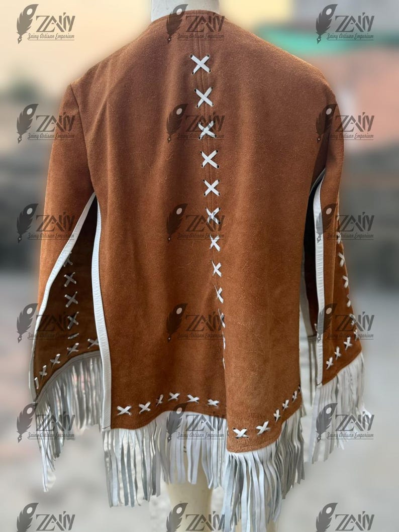 May include: A brown suede jacket with white fringe and accents. The back features white stitching in an X pattern. The jacket has a classic Western style with a bohemian flair. The sleeves and hem are adorned with long white fringe.