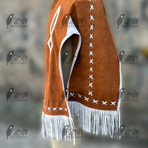 May include: A brown suede vest with white accents and fringe. The vest features white stitching and a geometric design on the front. The edges are trimmed with white leather and fringe. The vest is displayed on a mannequin.